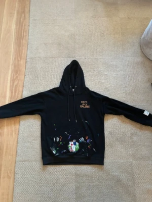 Svart hoodie med färgstänk och tryck - Säljer en svart hoodie med huva och dragsnören. På bröstet finns ett tryck med texten 'DEPT DE LA GALERIE' i guld. Nedtill syns färgstänk i flera färger både fram och bak. Hoodien har en avslappnad passform och ribbade muddar. Det är bara att ställa frågor om det skulle vara något 
