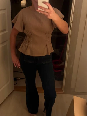 Beige blus med volang och kort ärm - Säljer en beige blus med korta, vida ärmar och volang nedtill. Toppen har smockdetaljer i midjan som ger en snygg form och är tillverkad i ett lätt, strukturerat tyg. Perfekt att styla med jeans eller kjol för en trendig look. Har även en i svart som jag säljer skriv vid intresse💞