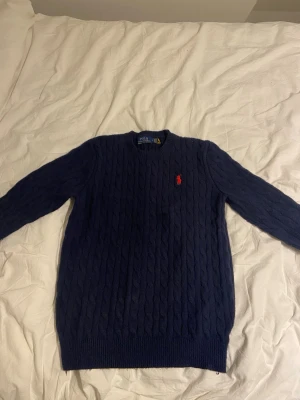 Mörkblå kabelstickad tröja Polo Ralph Lauren - Kabelstickad mörkblå tröja från Polo Ralph Lauren med klassisk röd logga broderad på bröstet. Tröjan har rund halsringning, långa ärmar och ribbade muddar. Perfekt för dig som gillar tidlös och stilren design. Den är L men den är liten i storlek så den e S/M
