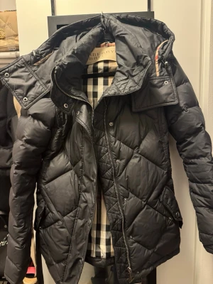 Svart dunjacka från Burberry Brit  - Snygg svart dunjacka från Burberry med klassiskt rutigt foder i beige, svart och rött. Jackan har huva, dragkedja och tryckknappar samt fickor med lock samt skärp i midjan. Perfekt för kalla vinterdagar och ger en exklusiv look.