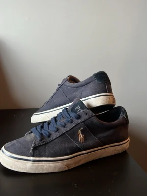  Polo Ralph Lauren Sneakers  - Snygga mörkblå sneakers från Polo Ralph Lauren med klassisk låg profil och vit sula. Skorna har blå skosnören, canvasmaterial och den ikoniska broderade loggan i guld på sidan. Perfekta för en avslappnad och trendig stil. Stolek 40