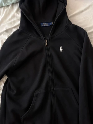 Svart hoodie från Polo Ralph Lauren - Klassisk svart hoodie från Polo Ralph Lauren med dragkedja framtill och vit broderad logga på bröstet. Hoodien har känguruficka och mjukt material som känns skönt mot huden. Perfekt för en avslappnad och stilren look.