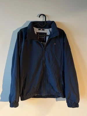 Höst jacka Tommy hilfiger  - Stilren mörkblå vindjacka från Tommy Hilfiger med dragkedja framtill, elastiska muddar och diskret logga på bröstet samt patch på ärmen. Jackan har randigt innerfoder och klassisk krage. Perfekt för blåsiga dagar. Storlek 164