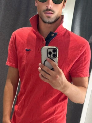 Röd polo vintage  Hollister - Röd polo från Hollister med klassisk krage och knappdetalj vid halsen. Polon har en liten mörkblå logga broderad på bröstet och är gjord i mjuk bomull. Perfekt för en avslappnad och stilren look. Står M men passar S 
