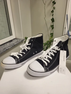Svarta high top sneakers med vita detaljer 44 - Snygga svarta high top sneakers från mywear med vita sulor, tåhättor och snören. Klassisk design med kontrastsömmar och metallöljetter på sidan. Perfekta för dig som gillar en stilren och tidlös look.
