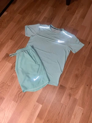 Mintgrönt träningsset från Nike - Snyggt mintgrönt träningsset från Nike med t-shirt och shorts. Båda plaggen har reflekterande Nike-loggor och är tillverkade i ett lätt, snabbtorkande material som passar perfekt för träning eller löpning. Passar storlek M också