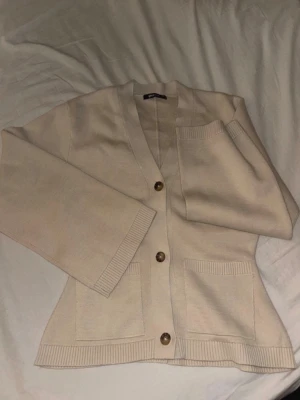 Beige kofta med knappar - Stilren beige kofta med v-ringning och stora bruna knappar framtill. Koftan har två rymliga fickor och ribbade muddar vid ärmslut och nederkant. Perfekt att slänga över en t-shirt för en avslappnad look. Helt oanvänd!