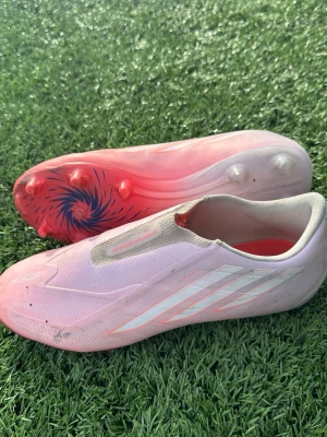 Rosa fotbollsskor utan snörning - Säljer ett par stilrena fotbollsskor i ljusrosa med vita detaljer och cool sula med mönster. Skorna har en modern design utan snörning och är perfekta för dig som vill sticka ut på planen. Yttermaterialet är syntet och sulan har fasta dobbar för gräs. ❗️Pris kan diskuteras ❗️