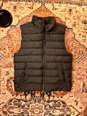Svart quiltad dunväst - Snygg svart dunväst med hög krage och dragkedja framtill. Västen har två praktiska fickor med dragkedja och en quiltad design som ger en modern look. Perfekt att slänga över en hoodie eller tröja för extra värme under kyliga dagar.