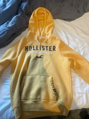 Gul hoodie från Hollister - Snygg gul hoodie från Hollister med trycket 'Hollister California' på bröstet och klassisk känguruficka. Mjuk insida, huva med dragsko och ribbade muddar. Perfekt för chill dagar och enkel att matcha med jeans eller joggers.