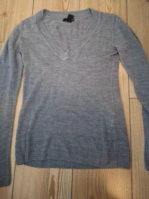 Grå stickad V-ringad tröja - En enkel och stilren grå stickad tröja med V-ringning och långa ärmar. Tröjan har ribbstickade muddar vid ärmslut och nederkant. Perfekt att matcha med jeans eller kjol för en avslappnad look.