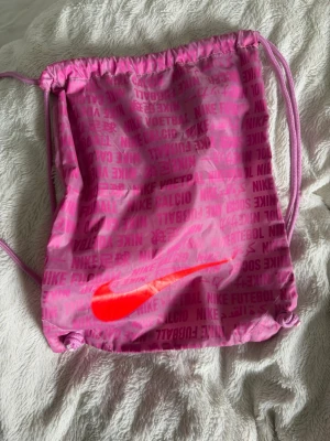 Rosa gympåse från Nike - Säljer en rosa gympåse från Nike med snörstängning och tryckt Nike-logga i neonrött på framsidan. Väskan har textmönster i olika språk och är gjord i ett lätt och slitstarkt syntetmaterial. Perfekt för träning eller att bära med sig det viktigaste.