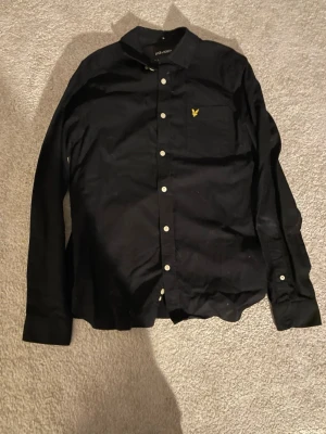 Svart skjorta från Lyle & Scott - Svart skjorta från Lyle & Scott med klassisk krage, vita knappar och lång ärm. Bröstficka med gul broderad logga. Skjortan är i bomull och har en stilren och tidlös look som passar till många tillfällen.