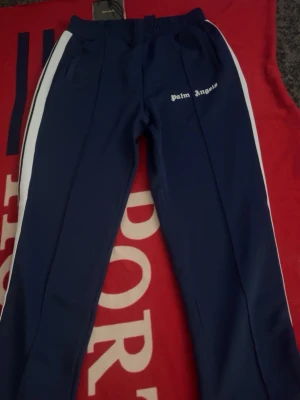Blåa Palm Angels mjukisbyxor - Säljer ett par blåa mjukisbyxor från Palm Angels med vita sidoränder och logga på vänster lår. Byxorna har resår i midjan och fickor på sidorna. Perfekta för en avslappnad och trendig streetwear-look.
