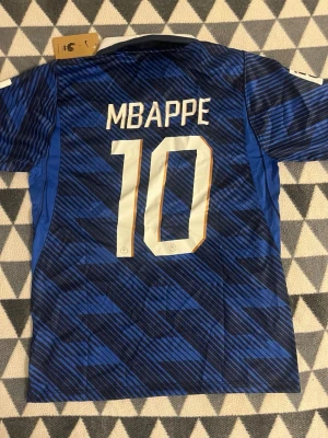 Blå Frankrike fotbollströja Mbappé 10 - Snygg blå fotbollströja med Mbappé och nummer 10 på ryggen. Tröjan har ett mönster i olika blå nyanser, vita detaljer och vit krage. Nike-logga och Frankrikes landslagsemblem på bröstet. Perfekt för fotbollsfans!