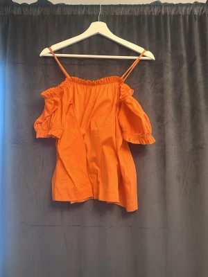 Orange offshoulder topp med volang - Trendig orange topp med offshoulder-design och smala axelband. Toppen har volangdetaljer vid ärmarna och en lös passform som ger en avslappnad look. Perfekt för varma dagar och enkel att styla med jeans eller kjol.