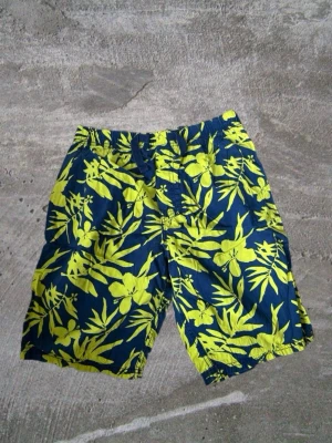 Blå-gula blommiga shorts  - Säljer ett par blå shorts med gult blommigt mönster. De har elastisk midja och är perfekta för sommaren eller stranden. Riktigt sköna och färgstarka shorts för dig som gillar att sticka ut.
