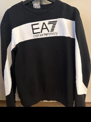 Svartvit sweatshirt från Emporio Armani - Snygg svart sweatshirt från Emporio Armani EA7 med vit panel över bröstet och armarna. Stor logga framtill. Klassisk rund halsringning och ribbade muddar. Perfekt för en trendig och sportig look.
