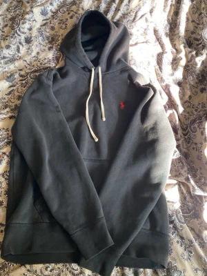 Svart hoodie från Polo Ralph Lauren - Klassisk svart hoodie från Polo Ralph Lauren med vit dragsko i huvan och röd broderad logga på bröstet. Hoodien har känguruficka framtill och är gjord i mjukt material som känns skönt mot huden. Perfekt för en avslappnad och stilren look.