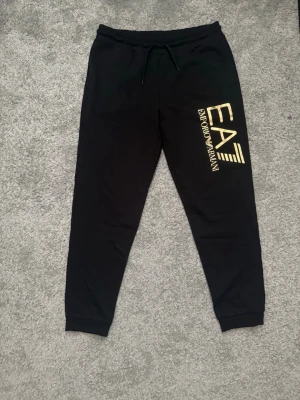 Svarta Armani EA7 mjukisbyxor  - Snygga svarta mjukisbyxor från Emporio Armani EA7 med guld logga på vänster ben. Byxorna har ALDRIG ANVÄNTS, alltså sjukt bra skick🤩.  Matrialet är kvalitet och det är skönt att ha på sig. Fraktar samma dag plagget är köpt!❤️‍🩹