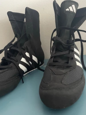 Svarta boxningsskor från Adidas - Säljer ett par svarta boxningsskor från Adidas med klassiska vita ränder på sidorna. Skorna har högt skaft, snörning hela vägen upp och slitstark sula. Materialet är en mix av mesh och syntet för bra ventilation och lätt känsla.