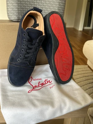 Mörkblå sneakers från Christian Louboutin - Snygga mörkblå sneakers i mocka från Christian Louboutin med klassisk röd sula och svarta skosnören. Insidan är beige och skon har en stilren, låg profil. Perfekt för dig som vill ha något exklusivt och trendigt.   FRAKTAS INOM 24 TIMMAR 