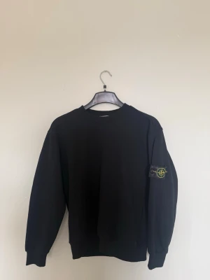 Svart sweatshirt  - Svart sweatshirt  med klassisk rund hals och ikonisk logotyp-patch på vänster ärm. Tröjan har ribbade muddar vid ärmslut och nederkant, perfekt för en clean och stilren look.