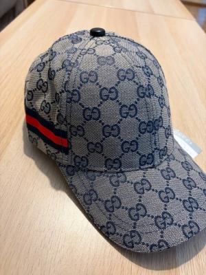 GG keps  - Snygg keps från Gg i beige tyg med mörkblått GG-monogram över hela kepsen. På sidan finns en röd och marinblå rand som ger extra stilpoäng. Klassisk böjd skärm och knapp på toppen. Perfekt accessoar för dig som gillar exklusiv streetwear.