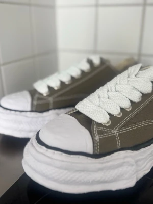 Maison Mihara Yasuhiro  - Snygga sneakers i olivgrönt tyg med vita, breda skosnören och kraftig, vit sula. Skorna har vita detaljer vid tån och hälen samt tydliga kontrastsömmar. Perfekta för dig som gillar en streetwear-look och vill sticka ut med en unik design.