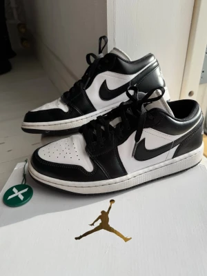 Svarta och vita Nike Air Jordan 1 Low skor - Snygga Air Jordan 1 Low sneakers i svart och vitt läder med klassisk swoosh på sidorna!🤩”Panda” modell. Skorna har lufthål uppe på tårna, svarta skosnören och låg siluett. Perfekta för dig som gillar streetwear och vill ha en ikonisk look🤍Dam Storlek 40 EU (25,5cm). Använda flera gånger men inga stora defekter eller några fel🙌Låda inkluderad. Köpta flera år sedan vid lansering på Stock X (ca 2000kr)! 100% äkta🩷Så fina skor som passar bra till alla plagg och sköna att ha på sig! Kan passa män. 