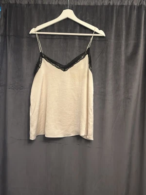 Beige linne med spetsdetaljer - Säljer ett stilrent beige linne med tunna axelband och svarta spetsdetaljer längs v-ringningen. Perfekt att styla lager på lager eller bära som det är för en trendig look. Materialet känns mjukt och lätt mot huden.