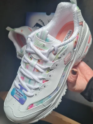 Helt nya vita Skechers sneakers med pastell - Snygga vita Skechers sneakers med färgglada pastellinslag i grönt, rosa och blått. Skorna har en chunky sula med mintgröna detaljer undertill och ovandel i syntetmaterial med meshpaneler. Klassisk snörning och mjuk memory foam-innersula för extra komfort. Helt nya och oanvända i kartong! Stl 38
