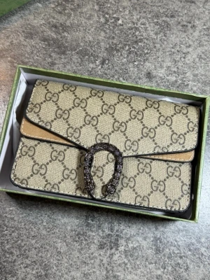 Beige GG kuvertväska  - Snygg beige kuvertväska med klassiskt GG-mönster över hela väskan. Väskan har en unik metallspänne framtill och är gjord i canvas med läderdetaljer. Perfekt för dig som vill ha en ikonisk accessoar med lyxig känsla.