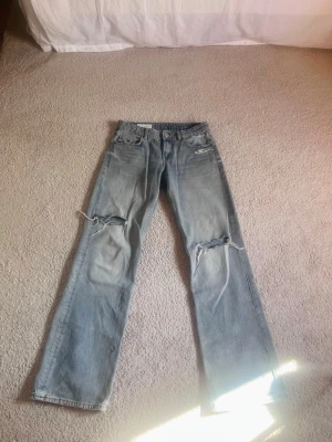 Low waist jeans med hål från ZARA - Säljer ett par ljusblå jeans från ZARA i low waist med raka ben och slitna detaljer över knäna. Jeansen har klassisk femficksdesign och är tillverkade i bomull. Perfekta för en avslappnad och trendig look.