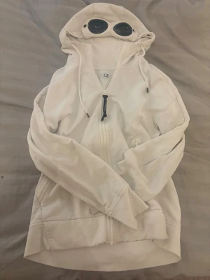 CP Company Hoodie  - Säljer en vit hoodie från C.P. Company med dragkedja framtill och ikoniska glasögon i huvan. Hoodien har fickor på sidorna, ribbade muddar och justerbar glasögon.