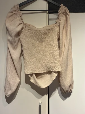 Beige blus med puffärmar - Trendig beige blus med smockad kropp och långa puffärmar. Fyrkantig ringning och rynkade detaljer vid axlarna ger en romantisk vibe. Perfekt att styla med jeans eller kjol för en snygg look.