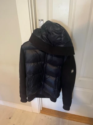 BVALDI cardigan  - Snygg svart pufferjacka med glansig framsida och stor vadderad huva. Ärmarna är i ett mjukare tyg och jackan har dragkedja framtill samt fickor. Perfekt för kalla vinterdagar och ger en sportig look.