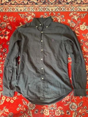 Svart skjorta från Ralph Lauren - Klassisk svart skjorta från Ralph Lauren med button-down krage och lila broderad logga på bröstet. Skjortan har långa ärmar, vita knappar och är tillverkad i mjuk bomull. Perfekt för en stilren och avslappnad look.