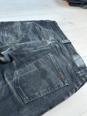 Svarta jeans från Nudie Jeans - Säljer ett par svarta jeans från Nudie Jeans med slitna detaljer och klassisk femficksmodell. Jeansen har raka ben och tydliga sömmar på bakfickorna. Materialet är denim i bomull och de har en cool, tvättad look!
