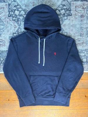 Mörkblå hoodie från Polo Ralph Lauren - Mörkblå hoodie från Polo Ralph Lauren med klassisk huva och vita dragsnören. Har en känguruficka framtill och den ikoniska röda Polo-loggan broderad på bröstet. Tillverkad i mjukt bomullsmaterial, perfekt för en avslappnad stil.