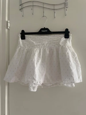Vit skort med broderade detaljer - Supersöt vit skort med volang och diskreta broderade prickar. Skorten har en bred resår i midjan och är gjord i ett lätt, luftigt tyg som känns perfekt för sommaren. Kjol framtill och shorts under gör den både snygg och bekväm.