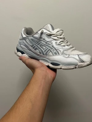 Vita och grå Asics sneakers - Säljer ett par vita och grå sneakers från Asics med klassisk mesh och syntetmaterial. Skorna har tydliga Asics-loggor på sidan och en chunky sula som ger retrovibbar. Perfekta för dig som gillar sportig stil och bekväma skor.