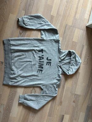 Grå hoodie från Gina Tricot - Säljer en grå hoodie från Gina Tricot med texten 'JE T'AIME' i svart på bröstet. Hoodien har huva och är tillverkad i mjukt, fleeceliknande material på insidan. Perfekt för en chill och avslappnad stil.