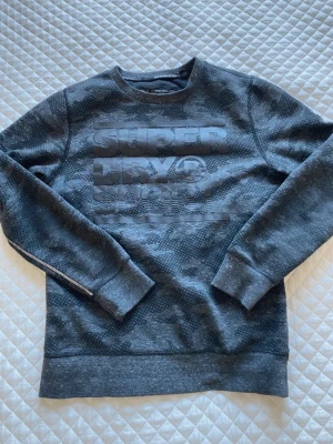 Mörkgrå sweatshirt från Superdry - Säljer en mörkgrå sweatshirt från Superdry med diskret kamouflagemönster och stor logga i relief på bröstet. Tröjan har rund halsringning, långa ärmar och ribbade muddar. Perfekt för en avslappnad och sportig stil.