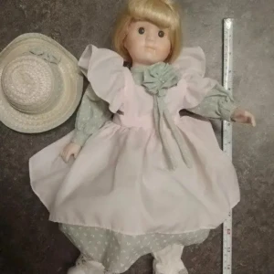 Söt porslinsdocka med ljus klänning - Charmig porslinsdocka med blont hår och stora bruna ögon. Dockan har en ljusrosa klänning med volanger, mintgröna byxor med vita prickar och matchande blus. Till dockan hör en ljus halmhatt med dekorativ rosett. Perfekt för samlare eller dekoration.