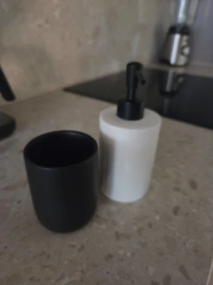 Svart mugg och vit tvålpump - Säljer ett set med en svart mugg och en vit tvålpump med svart pump. Båda är cylinderformade och har en stilren, modern design. Perfekt för badrummet eller köket. Materialet ser ut att vara keramik eller porslin.