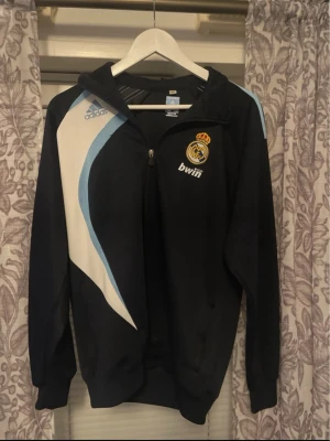 Real Madrid adidas träningsjacka - Snygg svart träningsjacka från adidas med Real Madrid-logga och Bwin-tryck på bröstet. Jackan har vita och ljusblå detaljer, dragkedja framtill och huva. Perfekt för fotbollsträning eller som sportig streetwear.