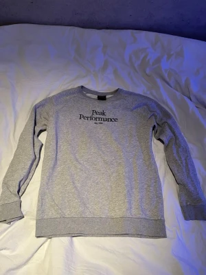 Grå sweatshirt Peak Performance - Snygg grå sweatshirt från Peak Performance med svart logga och texten 'Est. 1986' på bröstet. Tröjan har rund halsringning, långa ärmar och ribbade muddar vid ärmslut och nederkant. Perfekt för en avslappnad och sportig stil.