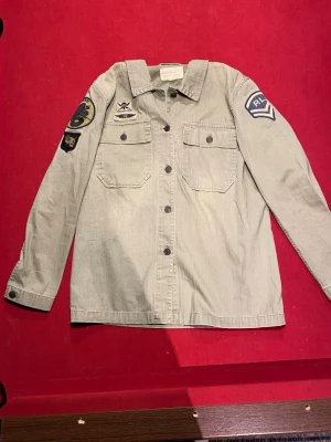 Grön overshirt från Ralph Lauren - Snygg militärgrön overshirt från Ralph Lauren med coola patchar på ärmarna och bröstet. Två bröstfickor med knappar och klassisk krage. Tillverkad i bomull och har en avslappnad passform som passar perfekt till streetwear-stilen.