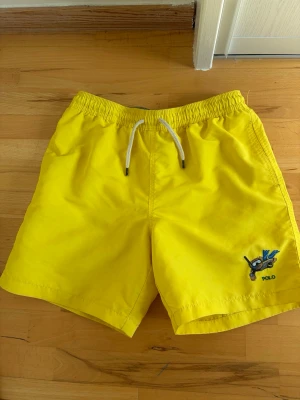 Gula badbyxor från Polo Ralph Lauren - Snygga gula badbyxor från Polo Ralph Lauren med broderad logga och motiv på ena benet. De har vit snörning i midjan och är tillverkade i polyester med meshfoder. Perfekta för sommaren och strandhäng!